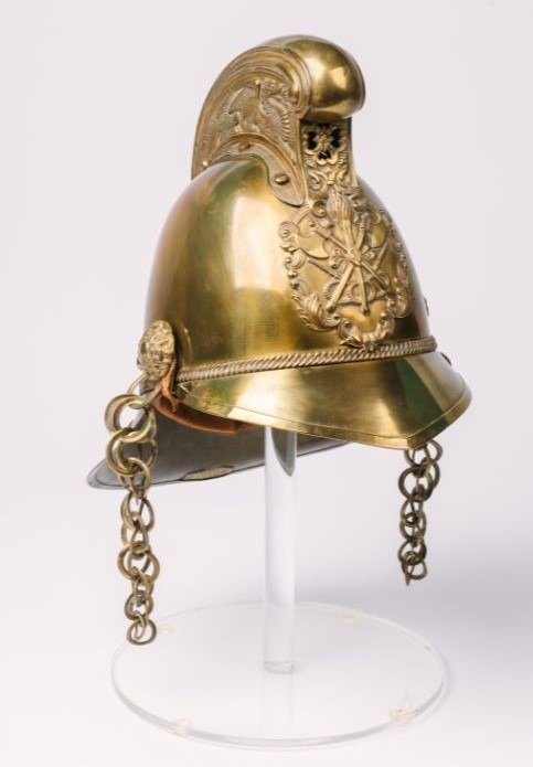 Image shows an elaborate brass helmet with chains on either side used to fasten it. The front badge has a design of fire, 2 axes and hoses. It has a Dragon design on both sides